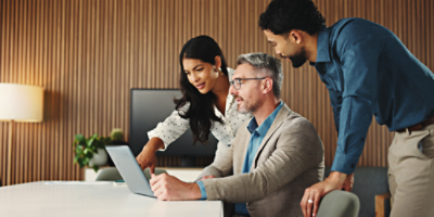 three people, one sitting and two standing over the shoulder looking at a laptop screen on a conference room meeting desk