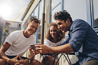 Three friends sitting outdoors, smiling and looking at a smartphone on a sunny day.
