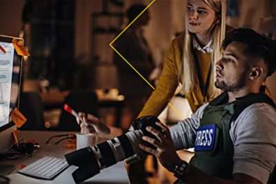 A journalist in a press vest holding a camera, working at a computer in an office with a colleague standing nearby.