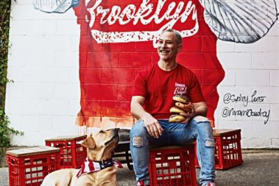 Brooklyn Boy Bagels owner Michael Shafran sitting on a red crate next to a dog in front of a large Brooklyn Boy Bagels mural 