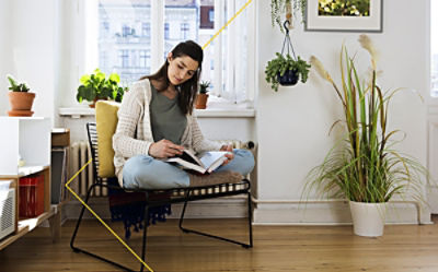 A young woman sits comfortably in a chair by a bright window, reading a book in a cosy, plant-filled room.