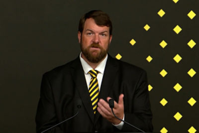 Man in black suit with a yellow and black stripped tie standing infront of a podium speaking into a microphone