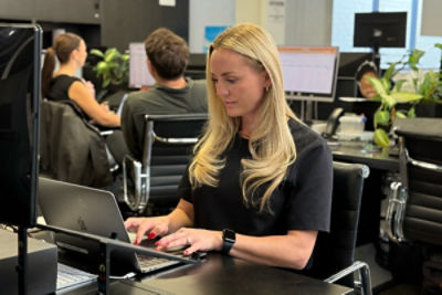 Women sitting at a desk in an office typing on a laptop with co-workers in the background talking