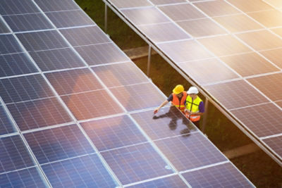 Large solar pannels in grids of four by eight. Two men in safety hard hat's and fluro vests standing between the panels inspecting them