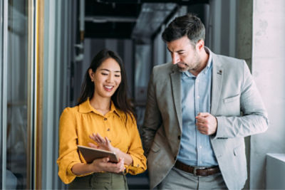 A woman on the left and a man on the right walking in an office looking at a tablet, discussing work