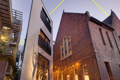 A modern white building beside a heritage red-brick church with stained-glass windows, illuminated by evening lights. A laneway perspective of Crystalbrook Albion boutique hotel in Sydney’s Surry Hills