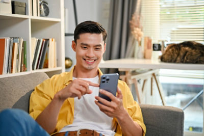Handsome and happy millennial Asian man in casual clothes using his smartphone, scrolling on social media while relaxing on sofa in his living room.