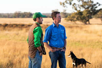 Two men and a cattle dog standing our in open farm land with tall dry grass