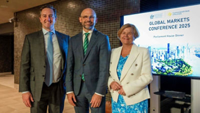 Three people standing having their photo taken infront of the sign for the global markets conference 2025